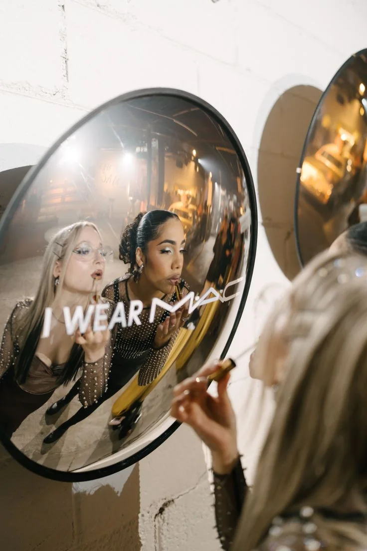 Two women applying lipstick in a round mirror